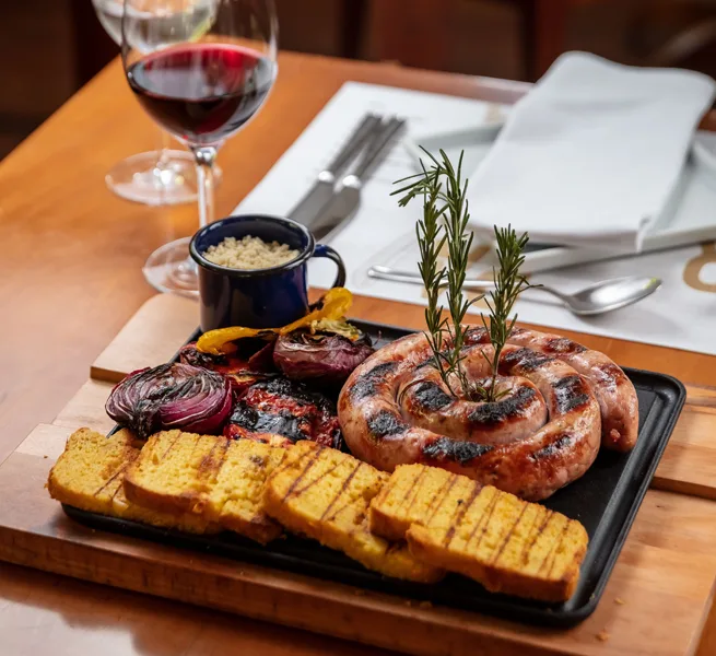 Linguiça em espiral com polenta grelhada e legumes, servida com vinho no ¡Vení! Parrilla