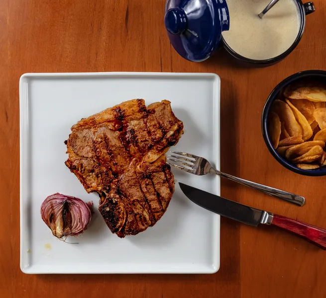 Corte T-bone grelhado com alho, batatas rústicas e molho, no ¡Vení! Parrilla