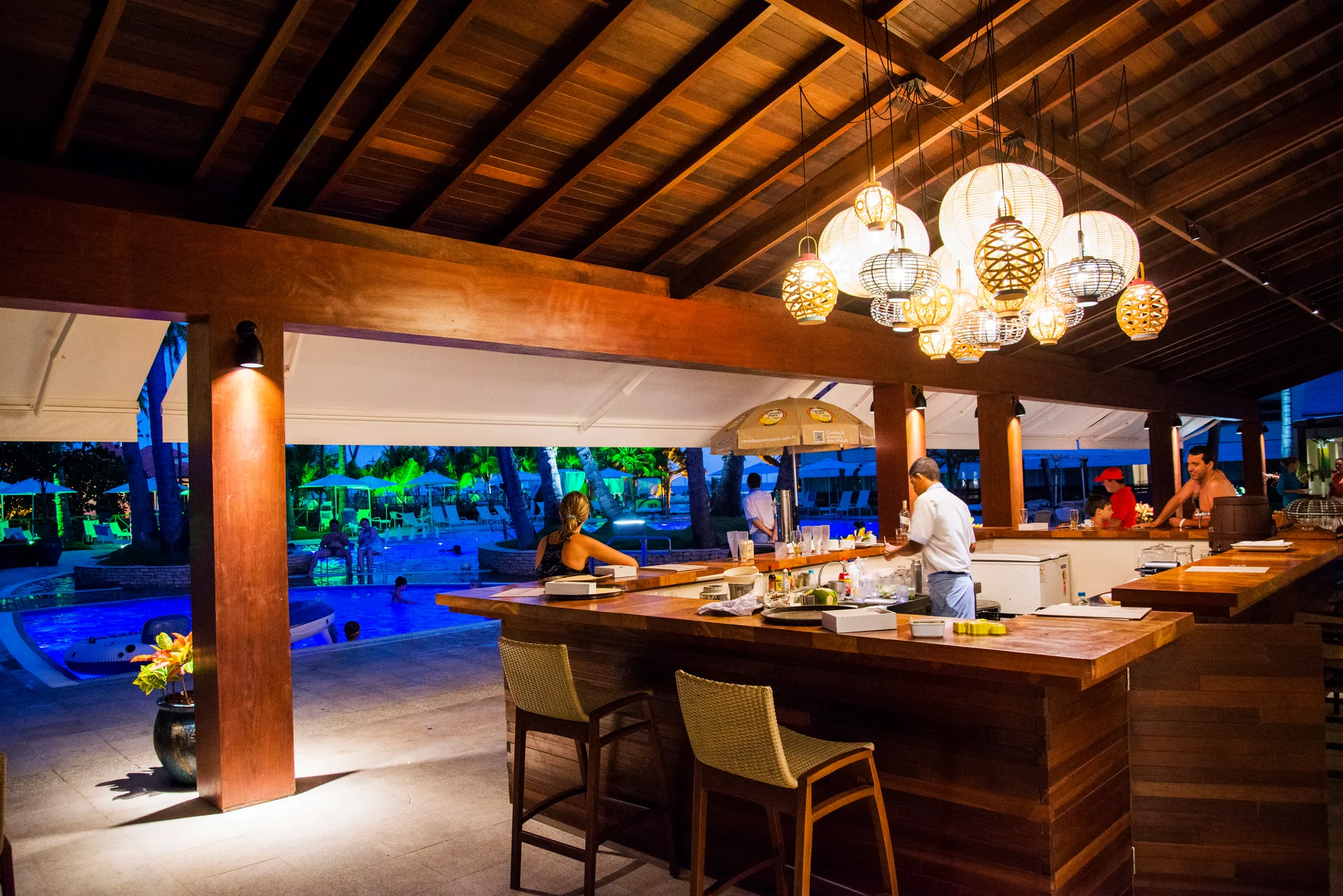 Jatiúca's Varanda Bar at dusk, wooden counter under lamps, with the pool lit up in the background