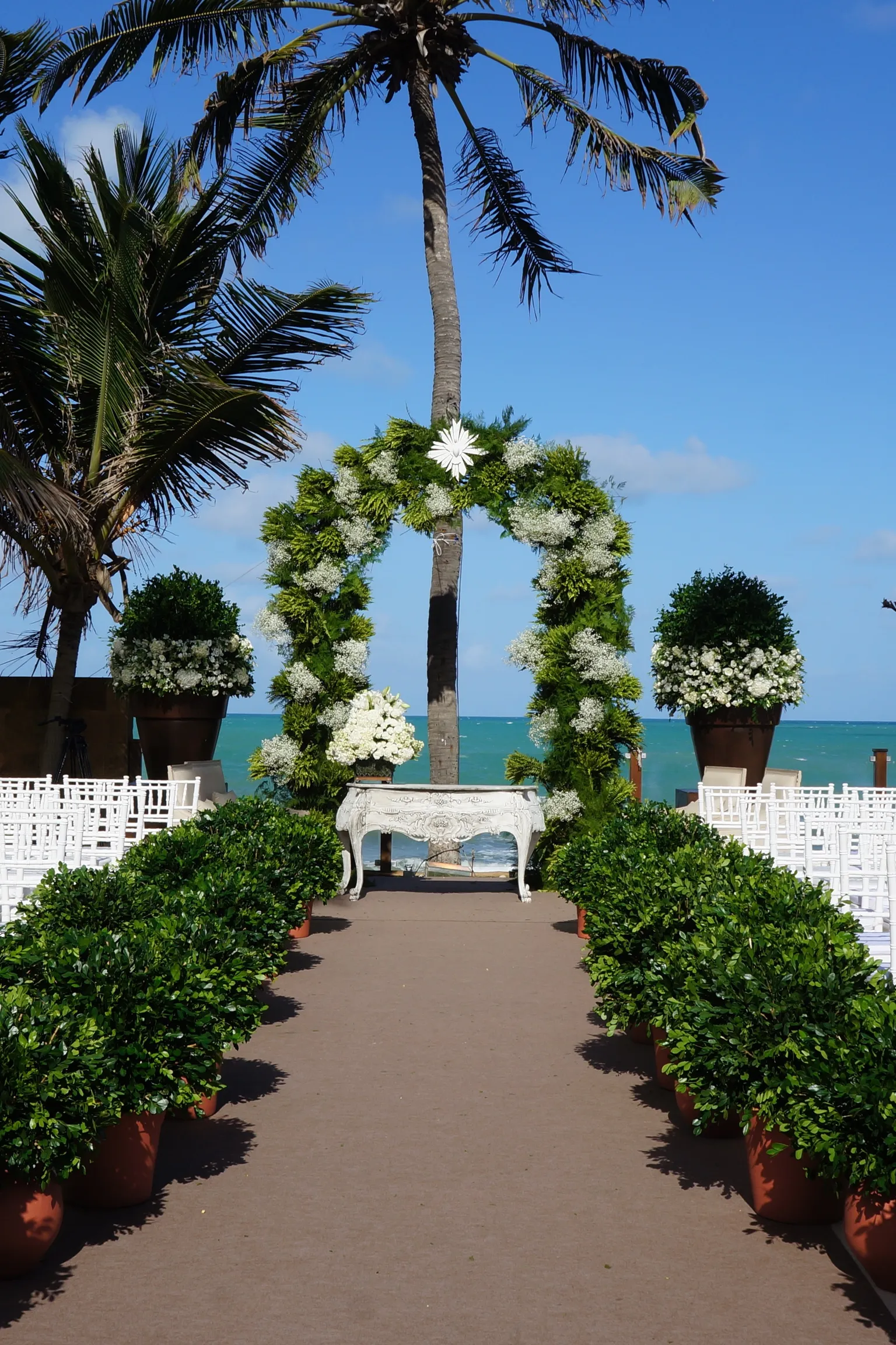 Cerimônia à beira-mar no Jatiúca, com corredor entre cadeiras brancas e arco de flores diante do mar